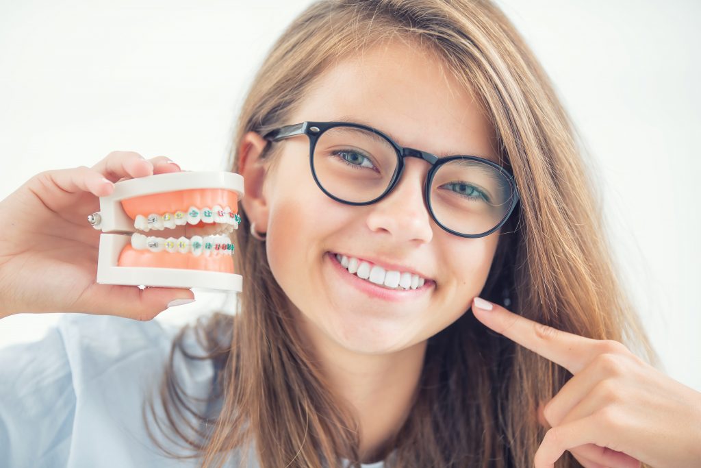 Young girl with glasses hold dental mol with braces while pointing to her newly straightened teeth.