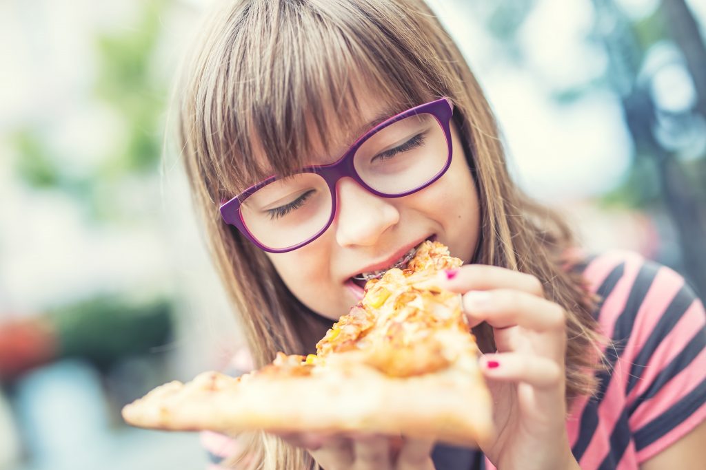 Little girl with purple glasses and braces eats a slice of cheese pizza