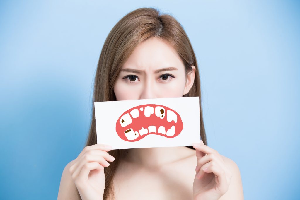 Braces & Tooth Decay: How to Avoid It & How to Spot It 1 Woman holds a paper cut out of an unhappy mouth featuring teeth with tooth decay in front of her mouth